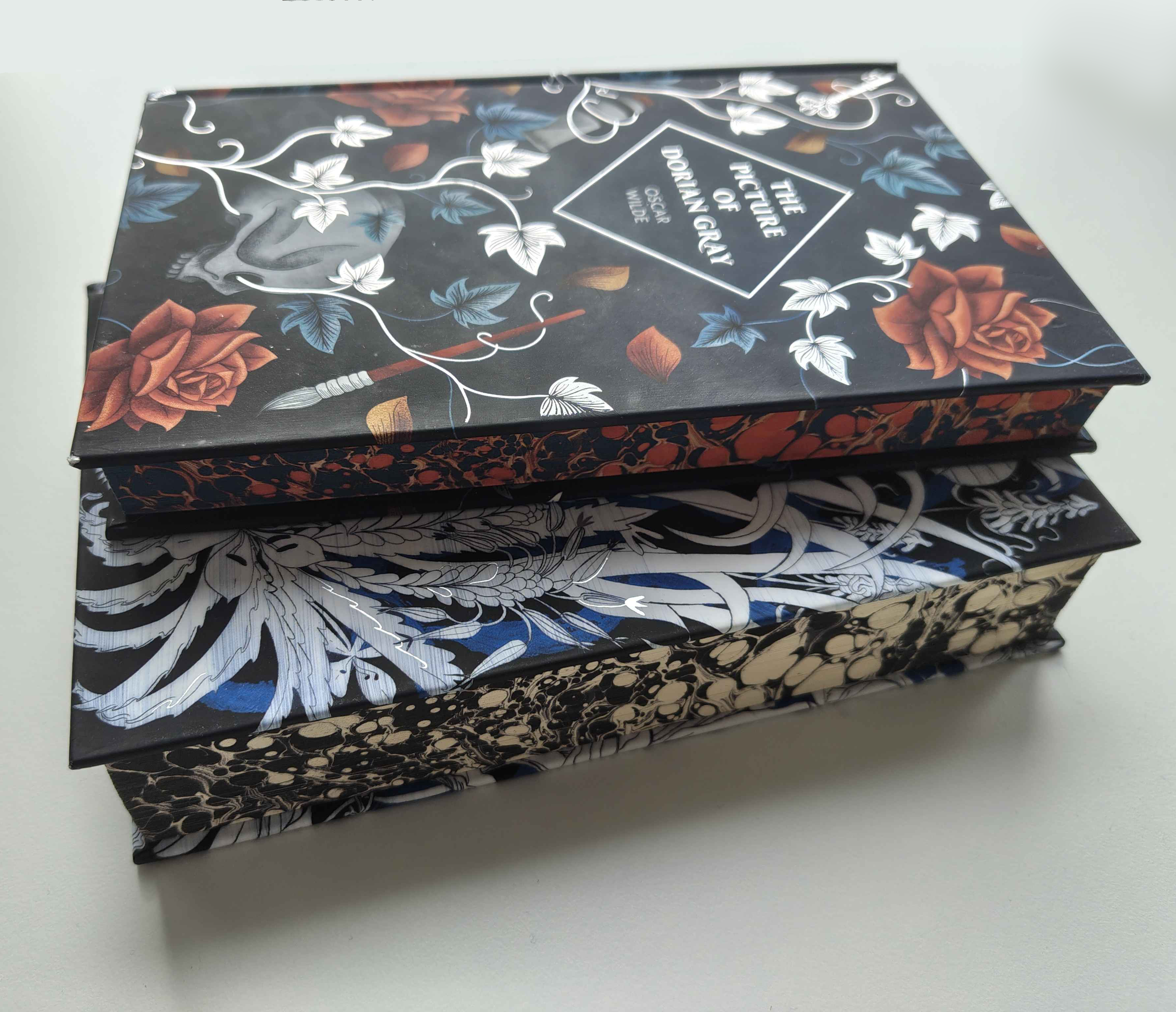 Two hard-backs stacked on top of each other, with marbled edges, and painted patterns representing themes of the book, some foiled. The top is The Picture of Dorian Gray, with red and blue marbling. The bottom is obscured, with white and black marbling.