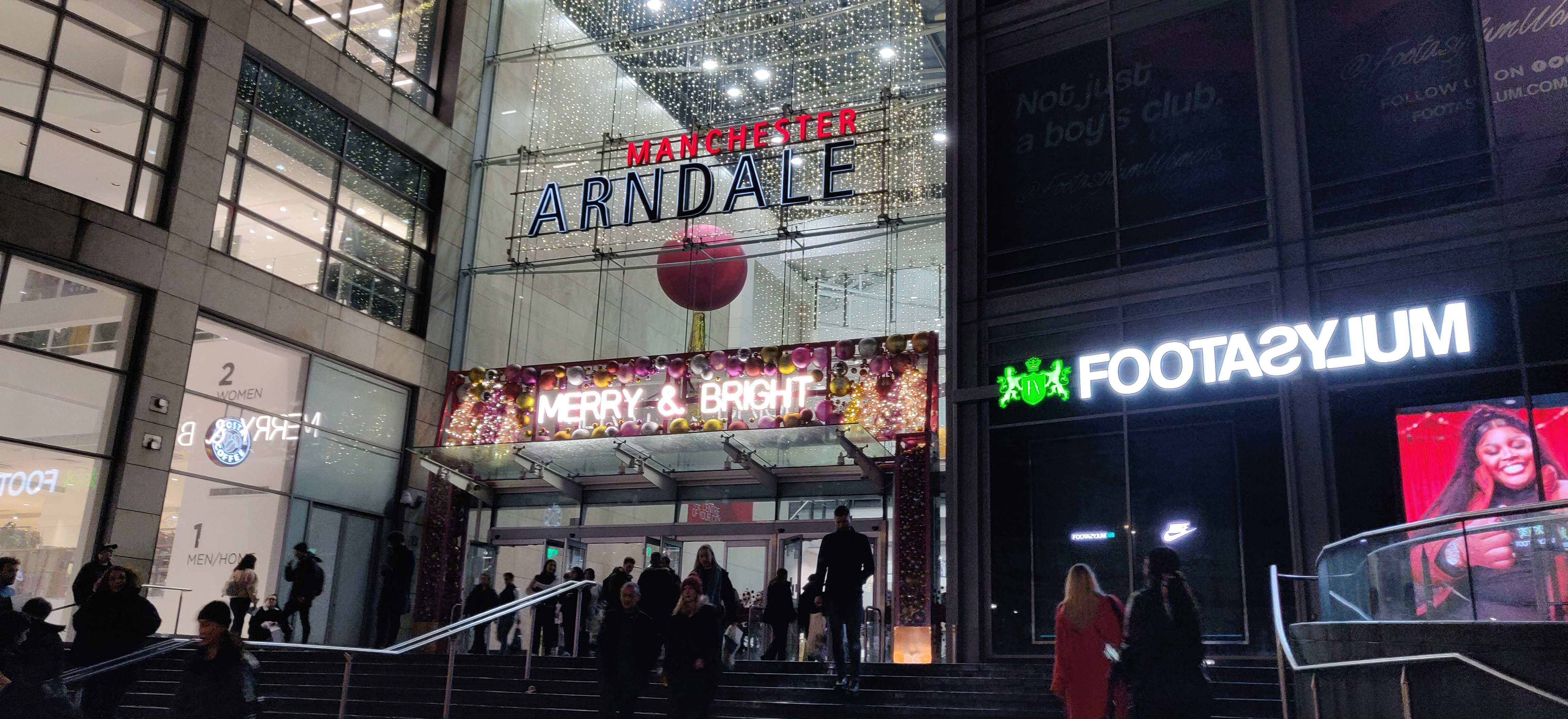 A photograph of the front of the Manchester Arndale, a shopping centre, at night. It’s Christmas and there are Christmas lights and decorations in the window. It’s busy with a crowd of people.