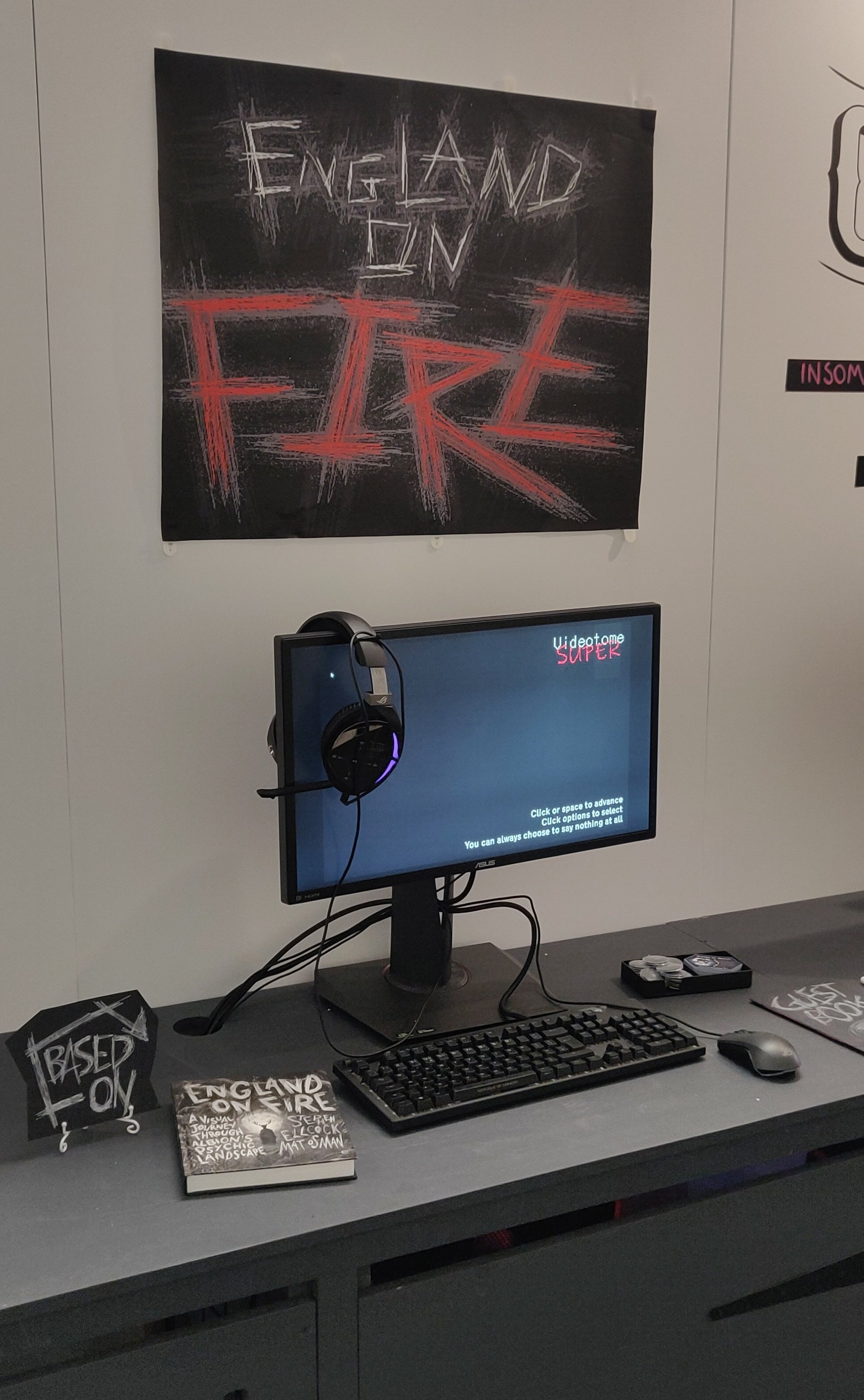 A convention booth setup with a PC playing ENGLAND ON FIRE. On the wall is a large poster with the game’s logo. On the desk is the original book, a pile of pins, and a guestbook.