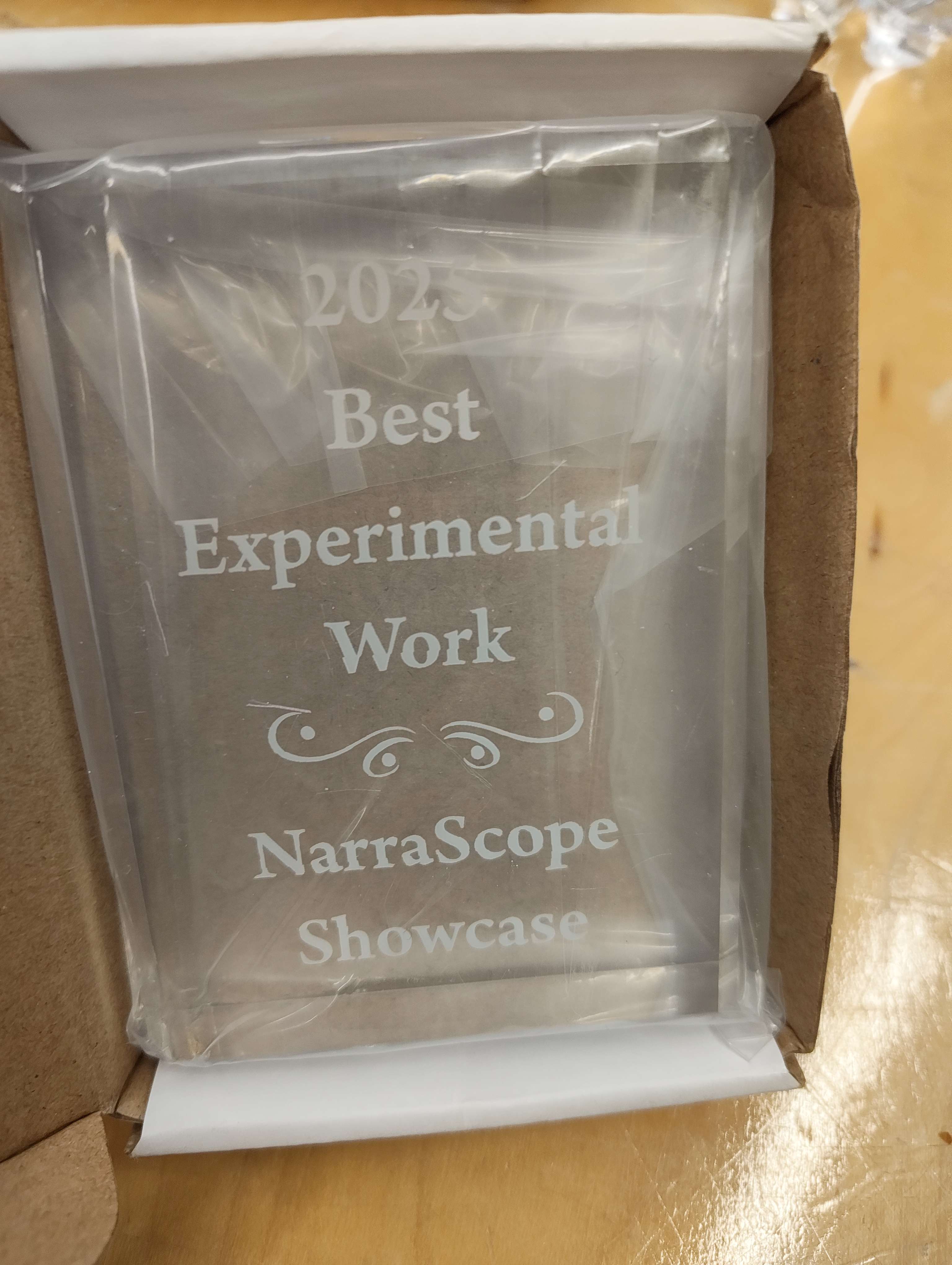 A photo of glass plaque wrapped in plastic, with text engraved on it. It reads: Best Experimental work, NarraScope Showcase